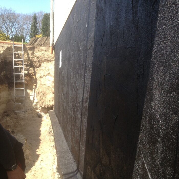 Blick auf eine schwarze Wand in einer Baugrube, mit einer Leiter und Sandboden im Vordergrund.