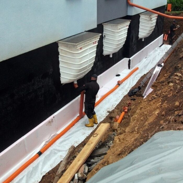 Arbeiter installieren Drainagesystem an einer Wand, mit Rohren und Eimer für Wasserableitung in einem Baubereich.